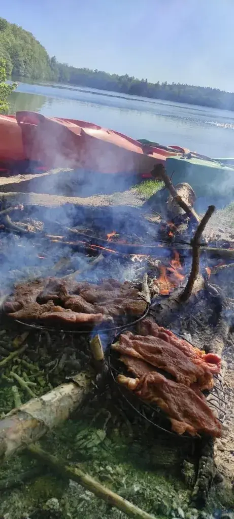 Mięso pieczone na ognisku nad brzegiem jeziora, w tle czerwone kajaki